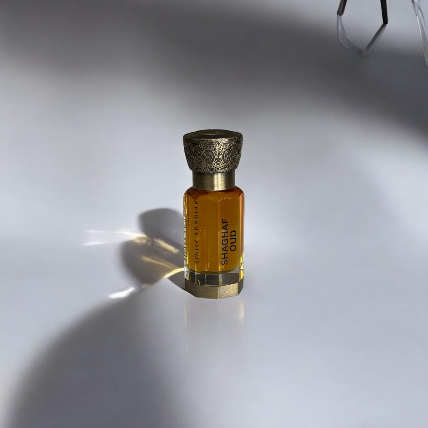 Flacon de parfum luxueux et compact, contenant un liquide ambré. L’étiquette affiche le nom 'SHAGHAF OUD' en lettres élégantes sur le verre transparent. Le bouchon doré, finement sculpté, ajoute une touche sophistiquée. L’éclairage subtil et l’ombre projetée sur la surface blanche mettent en valeur la brillance du flacon et l’intensité de la couleur du parfum.