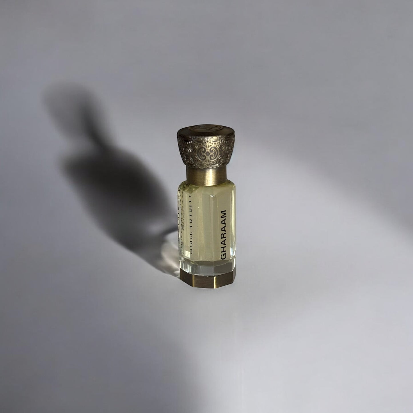 Flacon de parfum élégant et raffiné, en verre transparent contenant un liquide doré. Le nom 'GHARAM' est inscrit en lettres raffinées sur le côté du flacon. Le bouchon doré, finement gravé, apporte une touche sophistiquée. L’éclairage met en valeur l’ombre allongée du flacon sur un fond clair, ajoutant une dimension artistique à la présentation